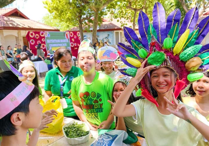 日本人の子どもが現地の子どもたちと一緒に、カラフルな野菜のかぶりものや手作りの帽子を身につけて、屋外で楽しそうに交流している様子。ELSAのSchooling Program中のアクティビティで、食育イベント「Nutrition Month」の一環と思われる。背景には装飾されたステージと「SPORTS FEST 2023」のTシャツを着た子どもも写っている。