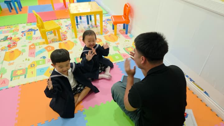 Global Standardのカラフルなマットの上で、先生と手遊びをする幼稚園児2人。