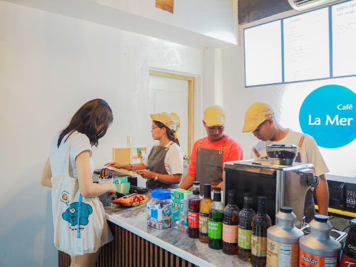 「Café La Mer」のロゴが掲げられた明るい校内カフェ。お揃いの帽子とエプロンを着用したスタッフが、コーヒーマシンや色とりどりのシロップが並ぶカウンターで生徒の注文に対応している様子。