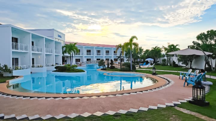 穏やかな夕暮れ時、ヤシの木と白いモダンな学生寮に囲まれた広大な屋外プール。プールサイドにはリラックスするための青いチェアや街灯が並び、リゾートのような落ち着いた雰囲気。