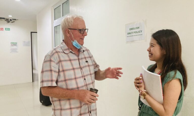 CLARK WE ACADEMYの講義棟で欧米ネイティブ講師と英語で話す留学生（廊下にはENGLISH ONLY POLICYの掲示）