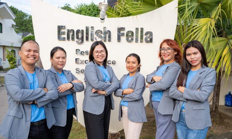 English Fella 2のヘッドティーチャー。学校のロゴの前に講師6人が並ぶ