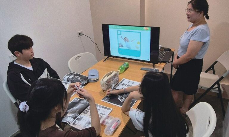 PIAのグループ授業。講師がモニターの前に立ち、留学生2人が教材を見ながら聞いている