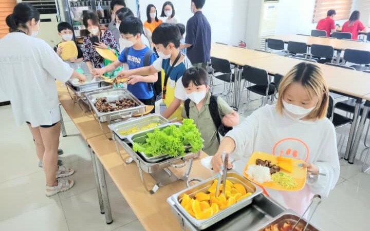 EG Academyの食堂でバイキング形式の昼食を楽しむ子どもたち。日本人や韓国人の学生が並び、栄養バランスの取れた料理が提供されている様子。