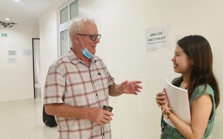 CLARK WE ACADEMYの講義棟で欧米ネイティブ講師と英語で話す留学生（廊下にはENGLISH ONLY POLICYの掲示）