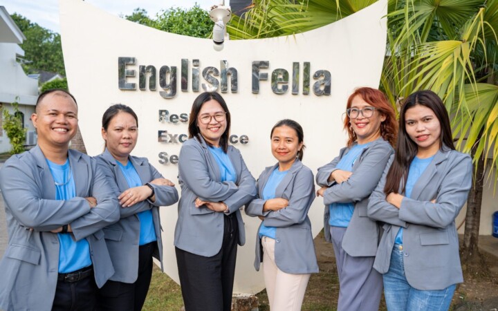 English Fella 2のヘッドティーチャー。学校のロゴの前に講師6人が並ぶ